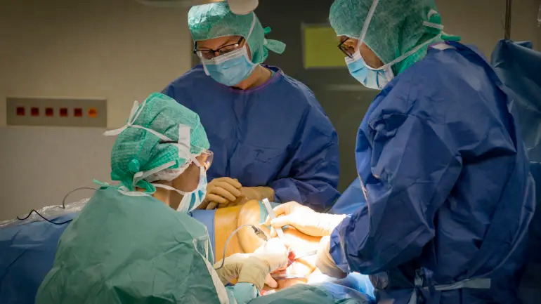 Três médicos especialistas em trajes cirúrgicos completos (toucas, máscaras, aventais azuis e verdes) estão concentrados realizando uma cirurgia em um paciente sob forte iluminação em uma sala de operação. Instrumentos cirúrgicos estão em uso sobre o paciente
