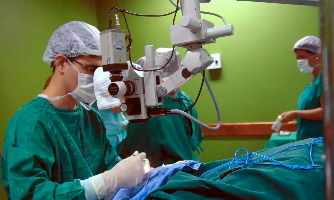 Em uma sala de cirurgia com paredes verdes, um profissional de saúde paramentado com touca, máscara, óculos e avental verde realiza um procedimento em um paciente coberto por campos cirúrgicos azuis, utilizando um microscópio cirúrgico. Ao fundo, outra profissional de saúde, também paramentada, auxilia.