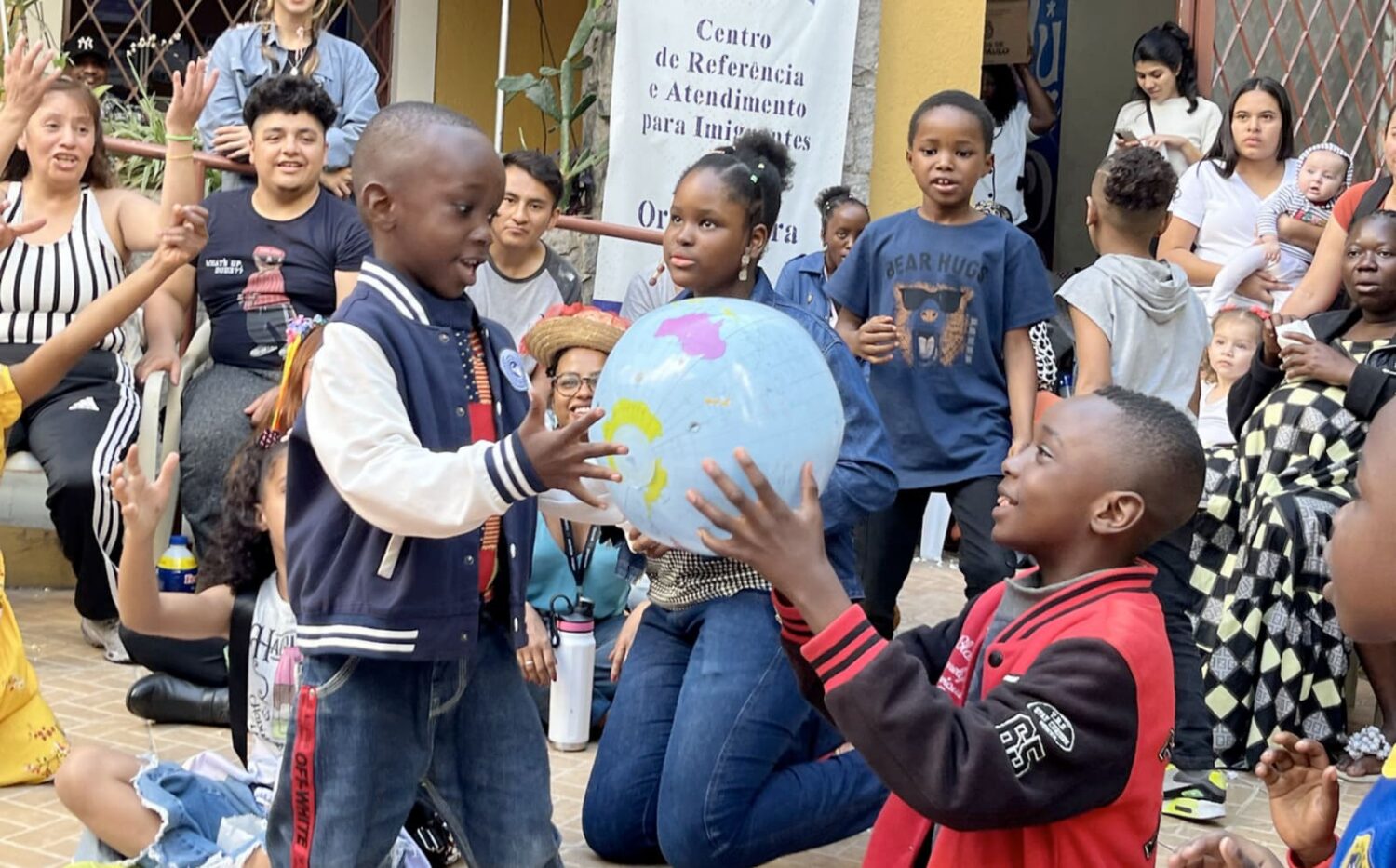 Crianças brincam com um globo inflável em uma atividade coletiva ao ar livre, cercadas por adultos e outras crianças. Ao fundo, há uma faixa do "Centro de Referência e Atendimento para Imigrantes". A cena mostra diversidade de origens e gerações reunidas em clima de integração e convivência.