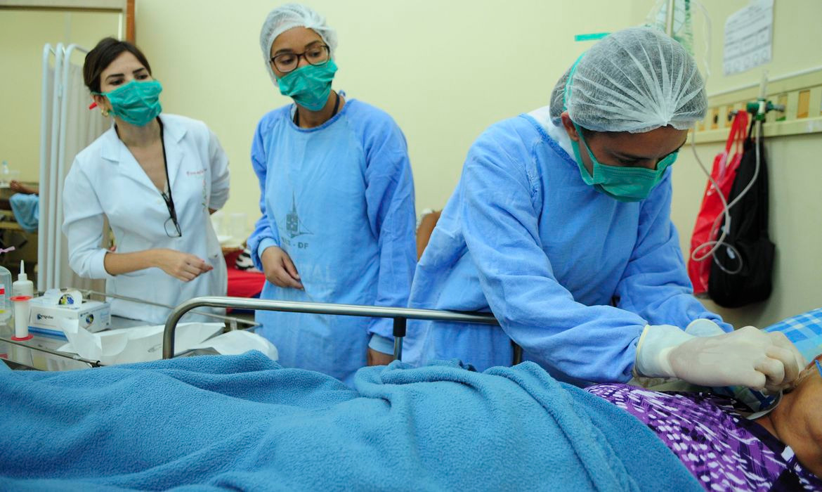 Três profissionais da enfermagem fazem o cuidado de uma pessoa em uma maca de hospital.