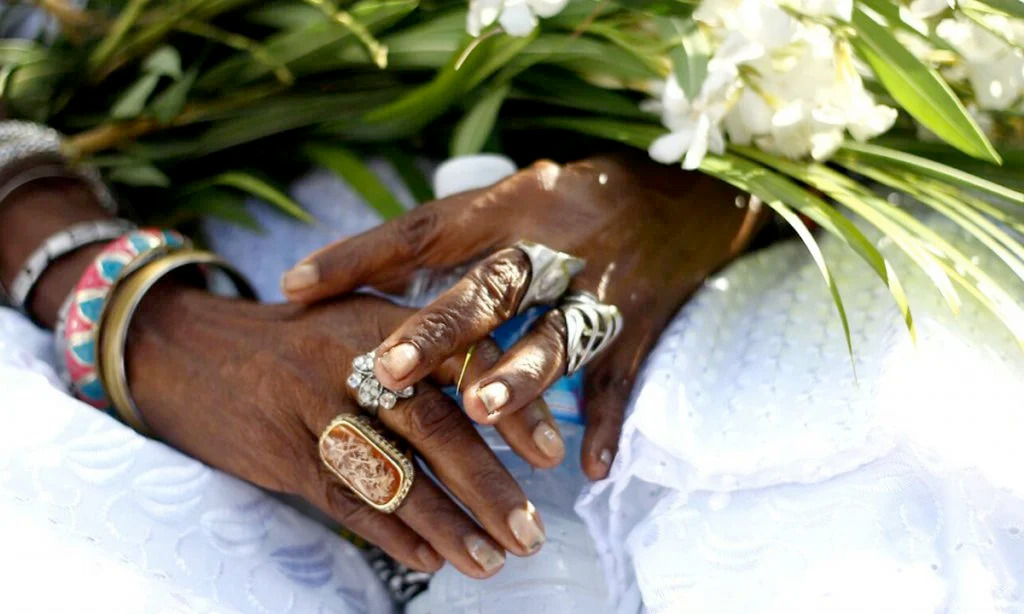 Mãos negras de uma mãe de santo, adornada com anéis e pulseiras e, sobre elas, um ramo de folhas e flores. Para combater racismo na saúde, é preciso valorizar a memória coletiva.